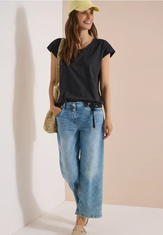 Een vrouw met een zwart t-shirt, wijde jeans, beige strotas en een pet tegen een lichte achtergrond.