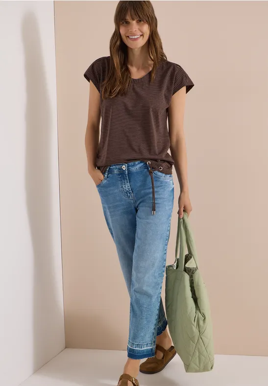 Frau vor hellem Hintergrund, trägt braunes T-Shirt, blaue Jeans und hält eine grüne Steppjacke.