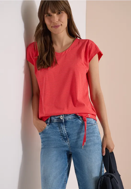Frau in rotem T-Shirt mit Knoten, blauen Jeans, hält dunkle Tasche, steht vor hellem Hintergrund.