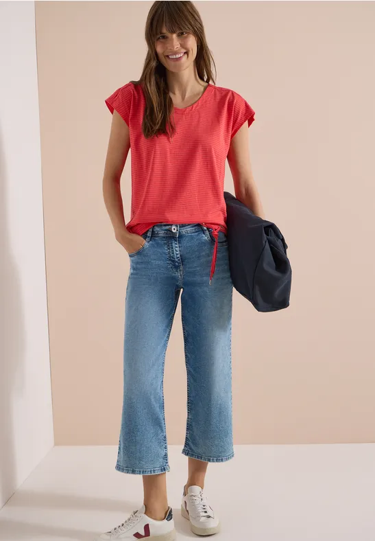 Frau in rotem T-Shirt, ausgestellten Jeans und weißen Sneakern, hält eine dunkelblaue Jacke.