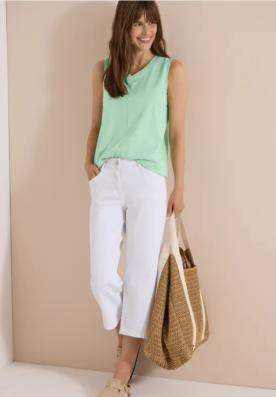 Frau in grüner Bluse, weißer Cropped-Jeans und Strohtasche vor beigem Hintergrund.