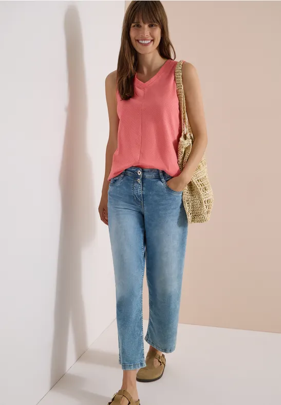 Frau in rosa ärmellosem Top, blauen Jeans, Basttasche und beigen Schuhen, stehend vor neutralem Hintergrund.