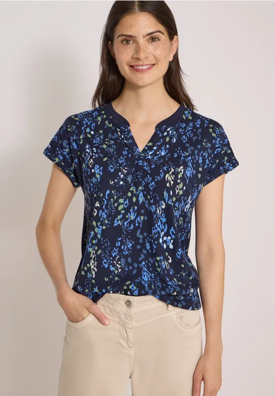 Frau mit marineblauer, kurzärmeliger Bluse mit Blumenmuster und beiger Hose vor hellem Hintergrund.