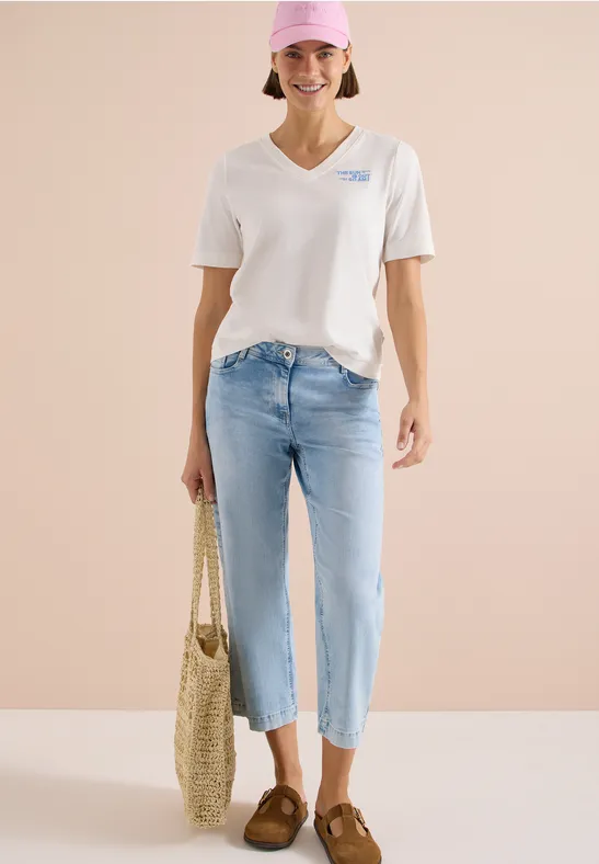 Frau in weißem V-Neck-Shirt, hellblauen Cropped-Jeans, braunen Loafers und Strohtasche vor beigem Hintergrund.