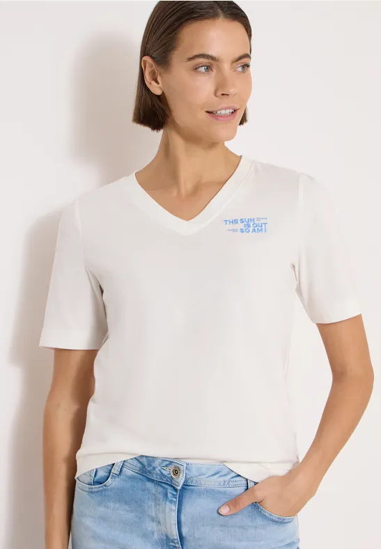 Frau in weißem V-Shirt und Jeans, eine Hand in der Hosentasche, vor hellem Hintergrund.