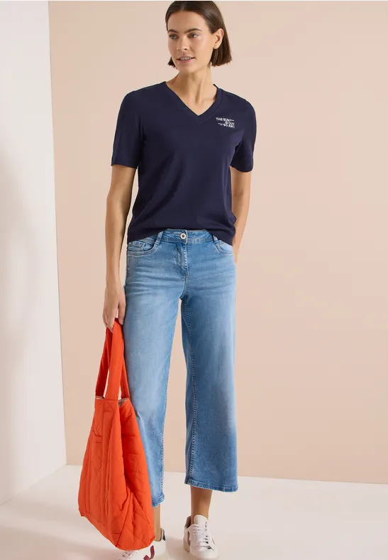 Frau in dunkelblauem Shirt, weitem hellblauen Jeans, weißen Sneakers und orangefarbener Tasche vor beigem Hintergrund