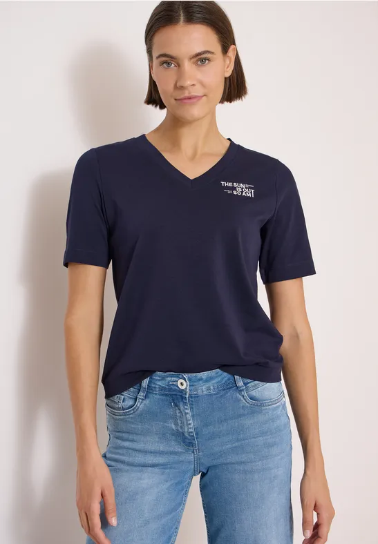 Frau in weißem T-Shirt und blauen Jeans vor hellem Hintergrund.