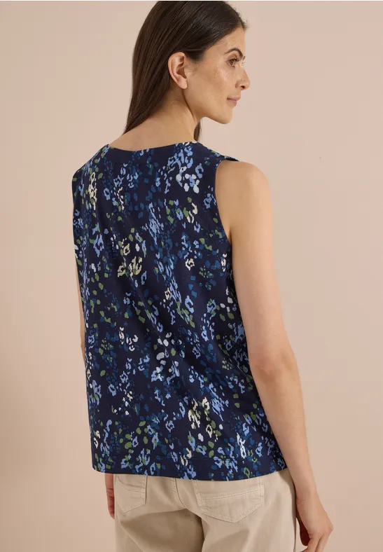 Frau von hinten im ärmellosen, dunkelblauen, floralen Top und beiger Hose vor hellem Hintergrund.