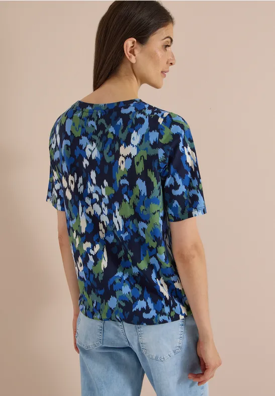 Une femme de profil porte un haut à manches courtes au motif floral abstrait bleu-vert et un jean clair.