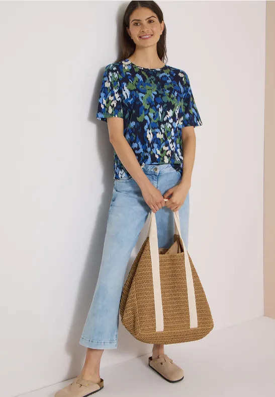 Femme en top floral bleu-vert, jean évasé clair, sac en paille et sabots beiges.