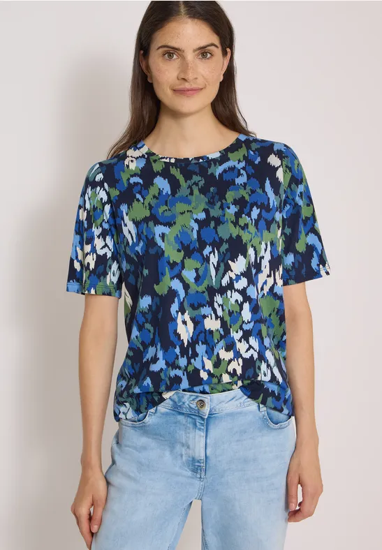 Femme portant un haut bleu à motifs verts et blancs avec un jean clair sur fond blanc.
