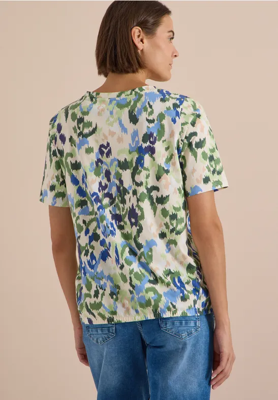 Frau von hinten mit kurzem Haar, gemustertem T-Shirt in Grün- und Blautönen und blauen Jeans.