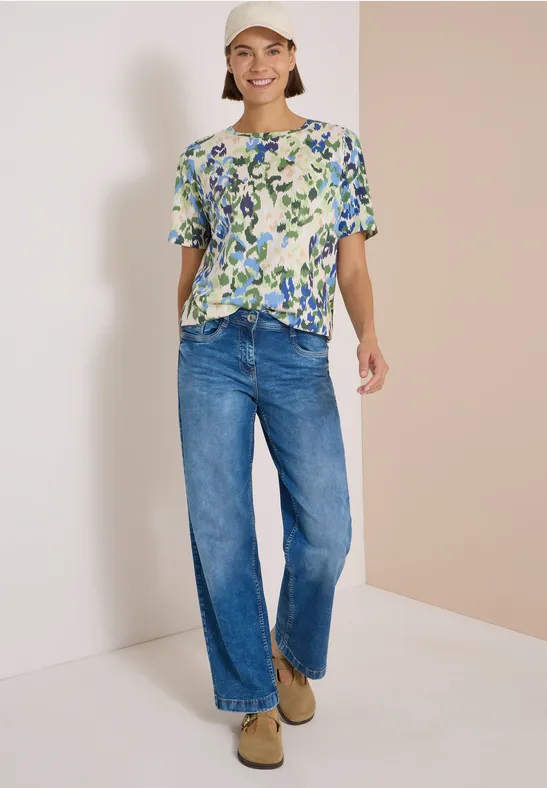 Dame trägt eine gemusterte Bluse mit Knopfleiste, weite Jeans und beige Schuhe, lächelnd vor neutralem Hintergrund.
