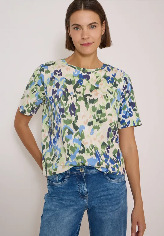 Frau trägt bunt gemustertes Kurzarmshirt mit grün-blauem abstraktem Blumenprint und Jeans.