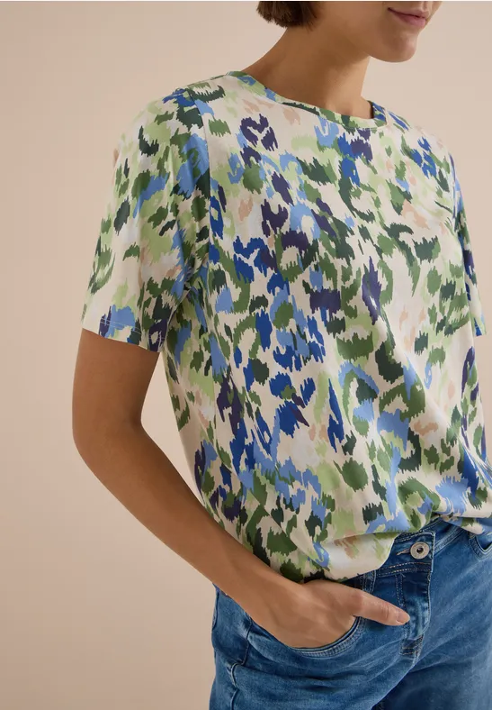 Frau trägt kurzärmlige Bluse mit abstraktem Blumenmuster in Blau und Grün und blaue Jeans.