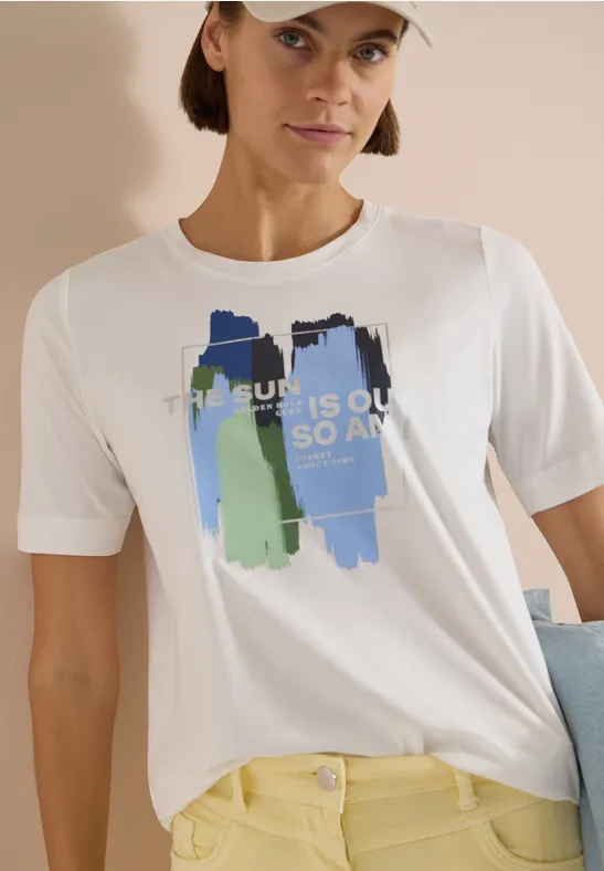 Wit T-shirt met blauw-groene verfstrepen en de tekst 'THE SUN IS OUT SO AM I', gecombineerd met een gele broek.