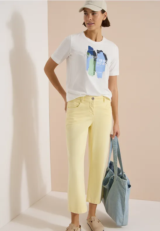 Vrouw in wit T-shirt met abstracte print, gele broek en blauwe jeanstas voor een lichte achtergrond.