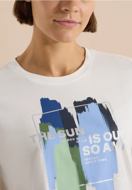 Vrouw met korte bob draagt wit T-shirt met abstracte penseelstreken in blauw en groen en een tekstprint.