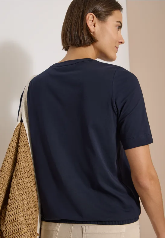 Frau mit dunkelblauem T-Shirt, beige Hose trägt Strohtasche, Rückenansicht.