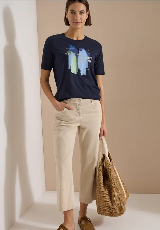 Frau trägt blaues T-Shirt mit abstraktem Print, helle Culotte und Korbtasche vor neutralem Hintergrund.