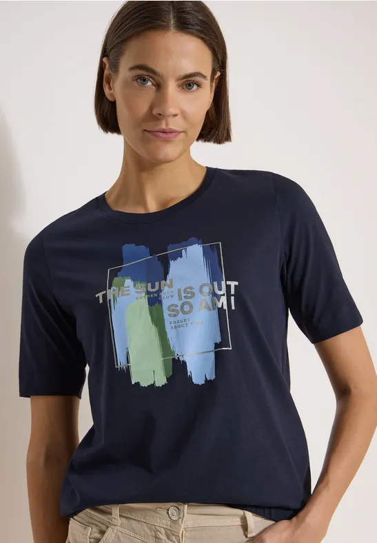 Person trägt dunkelblaues T-Shirt mit abstrakten Pinselstrichen in Blau und Grün und Schriftzug.