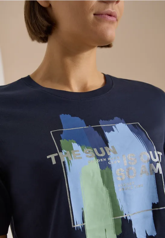Person trägt dunkelblaues T-Shirt mit abstrakten Pinselstrichen in Blau, Weiß und Grün und goldfarbenem Text.