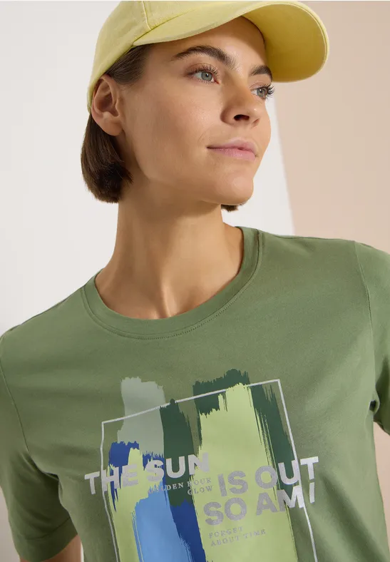 Person mit gelber Cap und grünem T-Shirt mit Pinselstrich-Design und Text