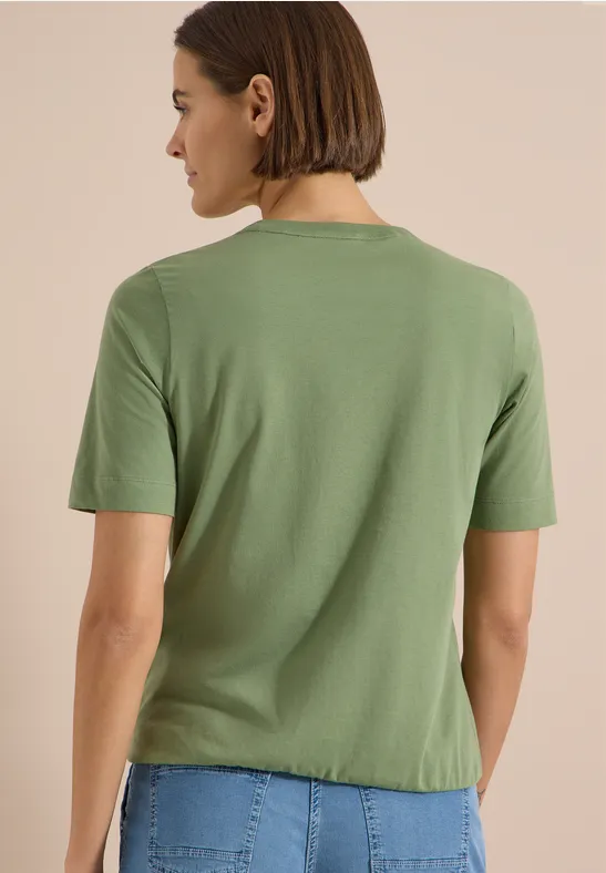 Frau von hinten in schlichtem grünem T-Shirt, kurze braune Haare, neutraler Hintergrund.