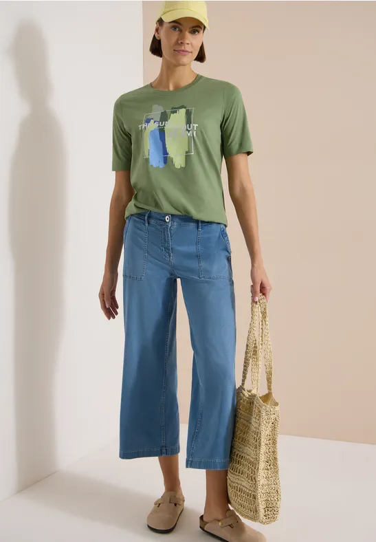 Person trägt grünes T-shirt mit Print, weite blaue Culottes, Basttasche und beige Loafers.