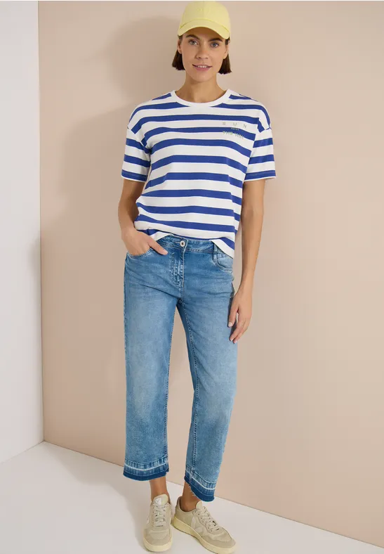 Vrouw met blauw-wit gestreept t-shirt, blauwe opgerolde jeans en witte sneakers tegen een neutrale achtergrond.