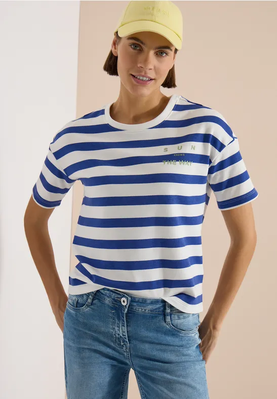 Persoon in wit-blauw gestreept T-shirt en spijkerbroek, handen in zij voor een neutrale achtergrond.