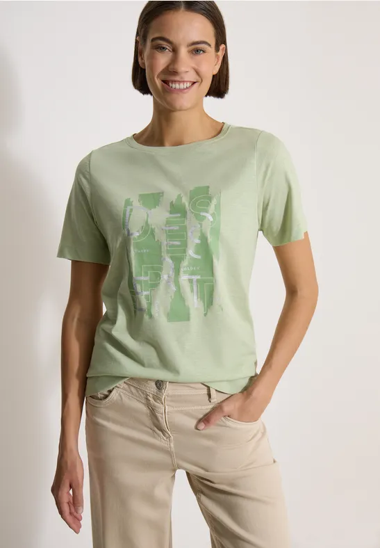 Frau trägt hellgrünes T-Shirt mit Grafikprint und beiger Hose, steht seitlich mit Hand in Tasche.