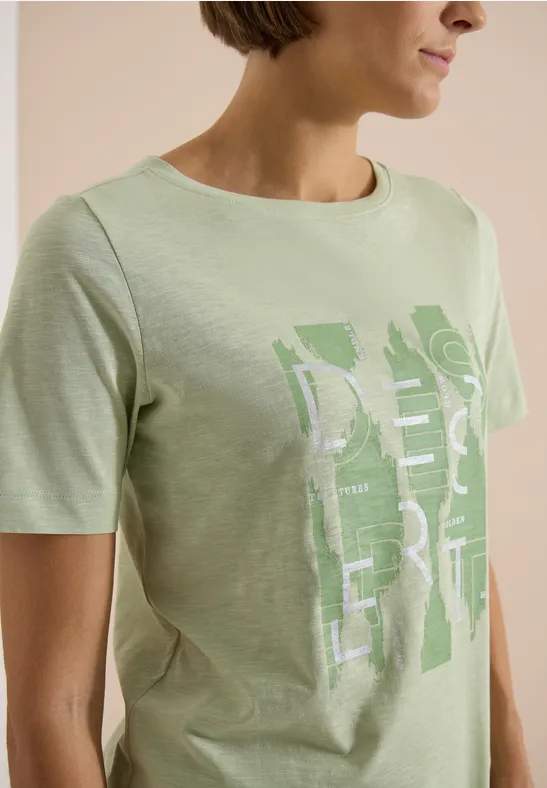 Frau in hellgrünem T-Shirt mit grafischem weißen/hellgrünen Print, vor schlichter beiger Hintergrund.