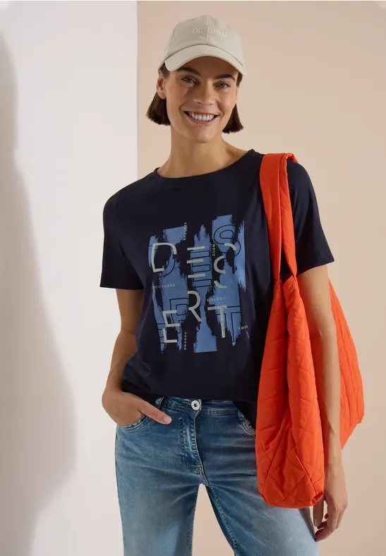 Person trägt Jeans, dunkelblaues Shirt mit Print, orange Umhängetasche und hellen Bucket Hat vor hellem Hintergrund.