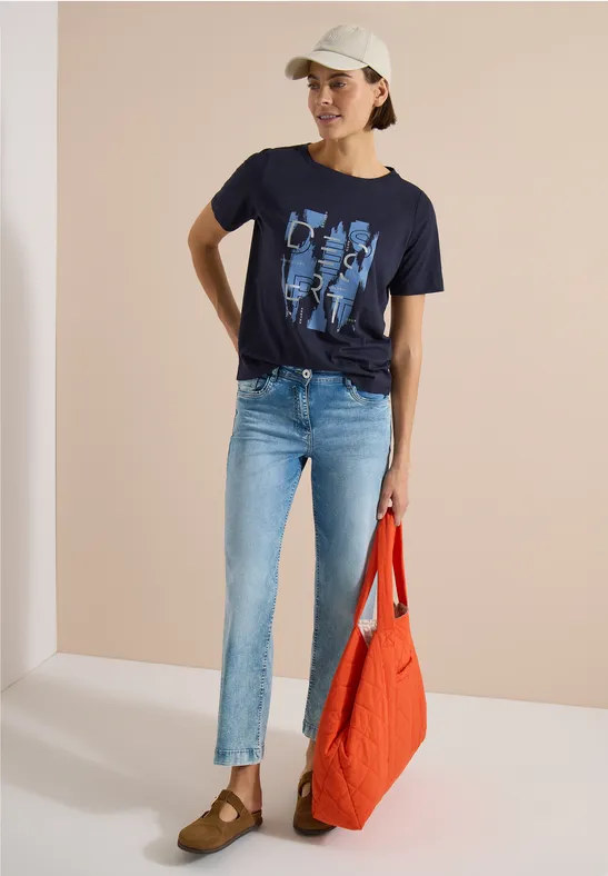 Frau in Jeans, dunkelblauem Shirt mit Print, Basecap und roter Tasche.