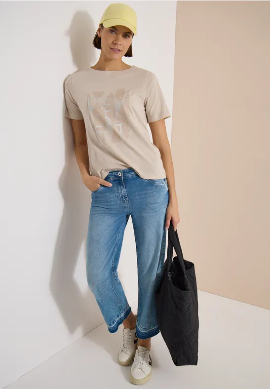 Person in beigem T-Shirt, blauen Jeans, weißen Sneakern und schwarzer Tasche.
