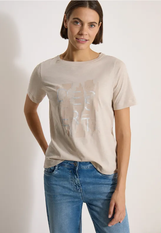 Frau in beigem T-Shirt mit hellem Aufdruck und blauen Jeans vor weißem Hintergrund.