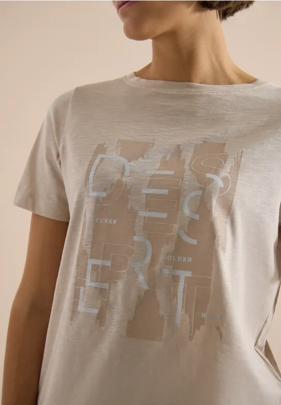 Eine Frau trägt ein beiges T-Shirt mit abstraktem, silbernem Schriftzug