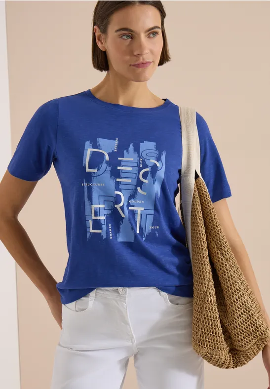 Frau trägt blaues T-Shirt mit 'Desert'-Print, weiße Hose und große Strohtasche über der Schulter