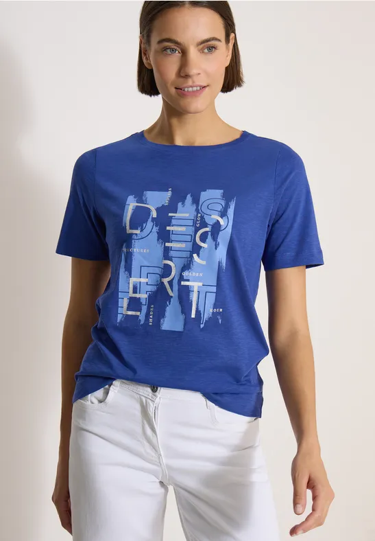 Person trägt blaues T-Shirt mit abstraktem Schriftzug und weißer Hose, steht entspannt vor hellem Hintergrund.