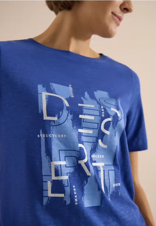 Frau trägt blaues T-Shirt mit großem, abstraktem Schriftzug 'DESERT' in hellem Kontrast.