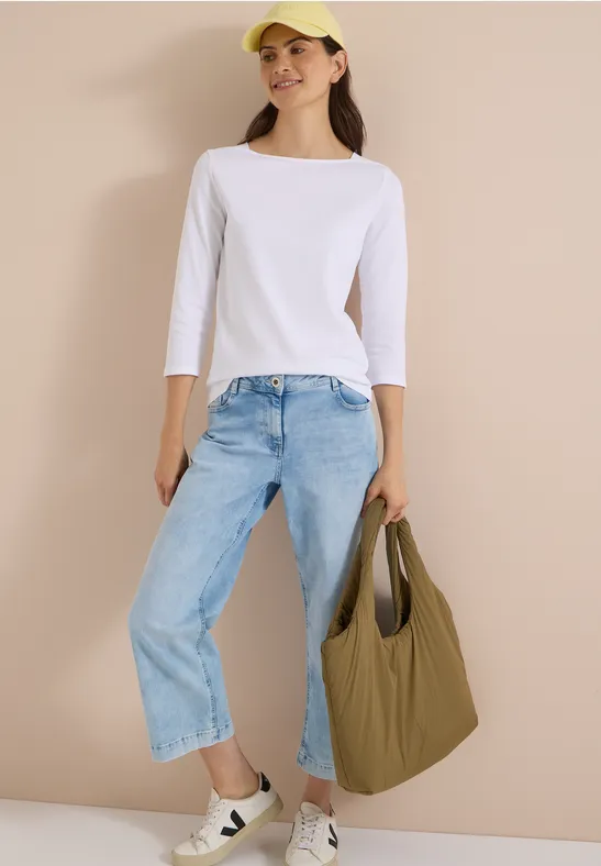 Frau in weißem ¾-Arm-Shirt, hellblauen Jeans und gelber Cap trägt eine khakifarbene Tasche.