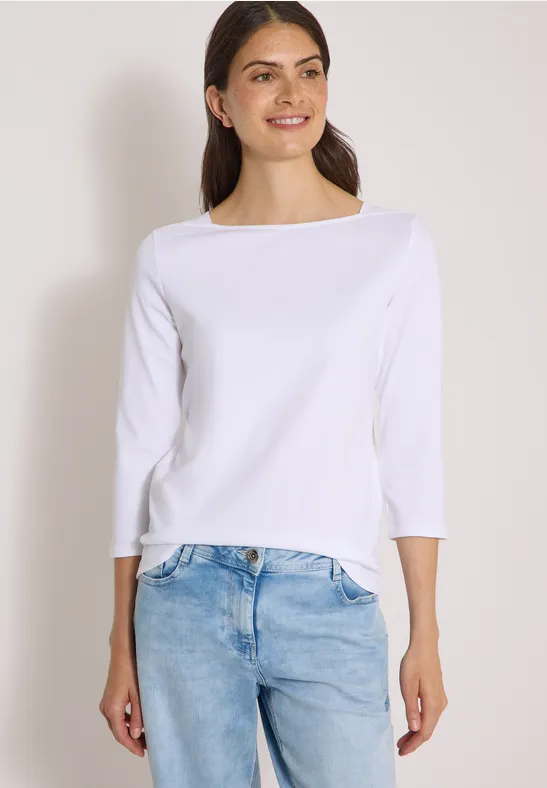 Frau in weißem Dreiviertelarm-Shirt und hellblauen Jeans lächelt seitlich vor hellem Hintergrund.