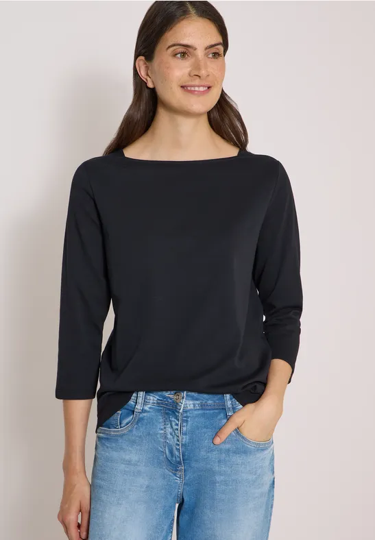 Junge Frau in schwarzer 3/4-Ärmel-Bluse mit Boot-Ausschnitt und blauen Jeans, lächelnd mit Hand in Tasche