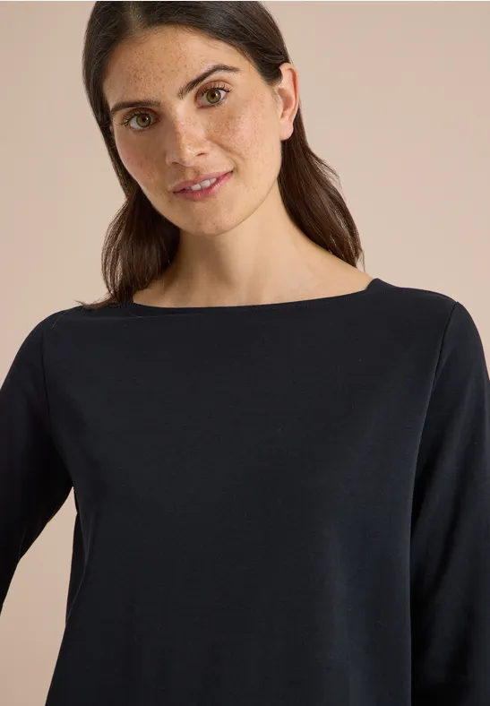 Porträt einer lächelnden Frau mit dunklem Langarmshirt vor neutralem Hintergrund.