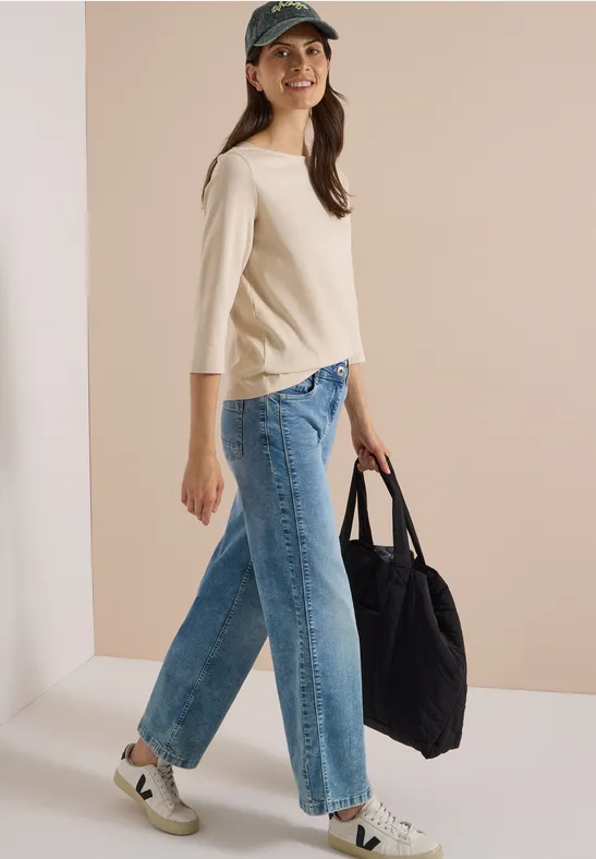 Frau mit Cap, hellbeigem Shirt, weiten Jeans und Sneakers geht lächelnd, trägt eine große schwarze Tasche.