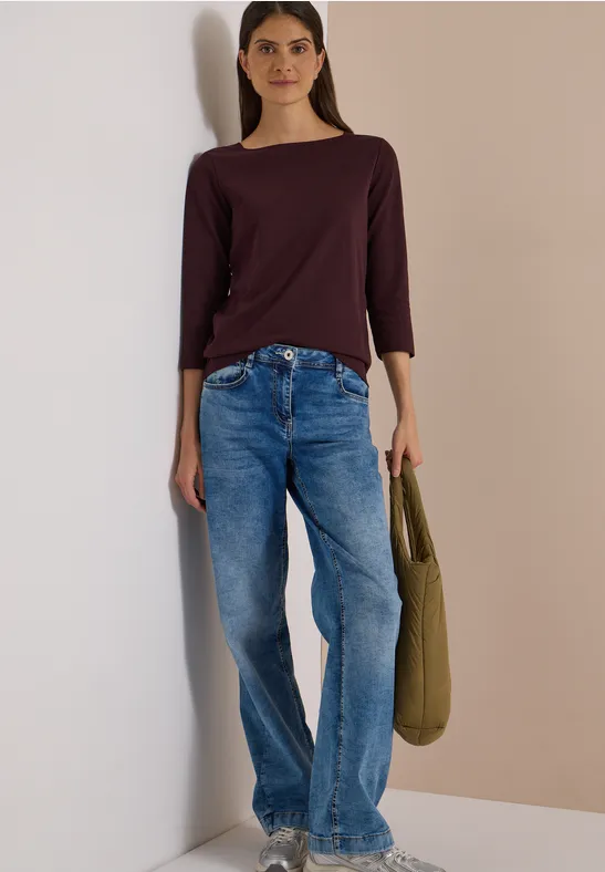 Frau steht vor einer Wand, trägt bordeauxfarbenes Shirt, blaue Jeans und eine grüne Tasche.