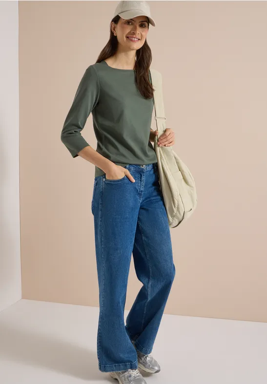 Frau in grünem Langarmshirt, weiten Jeans, Sneakern, weißer Cap und Beuteltasche vor neutralem Hintergrund.
