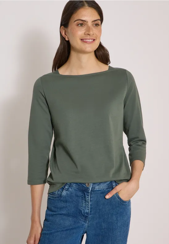 Frau mit grünem Langarmshirt und blauen Jeans lächelt, Hand in Tasche.