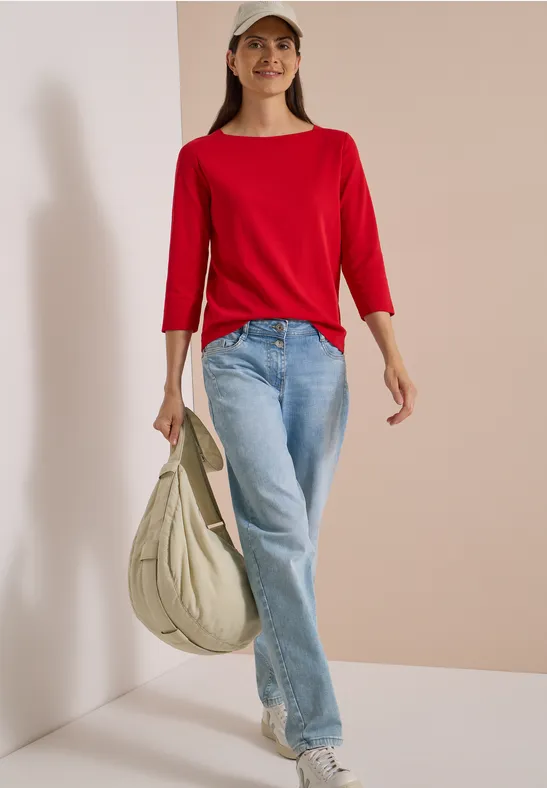 Frau in rotem Shirt, Jeans, weißen Sneakern und Cap mit großer beiger Tasche vor neutralem Hintergrund.